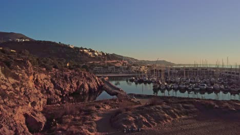 Port-ginesta-at-sunrise-with-boats-docked-and-cliffs-in-the-foreground,-aerial-view