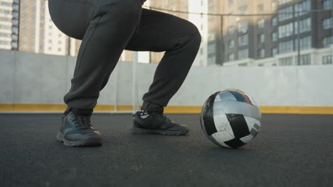 lower angle view of man in squatting position with hands clasped, performing intense workout on outdoor sports ground with soccer ball rolling on ground and urban building in background