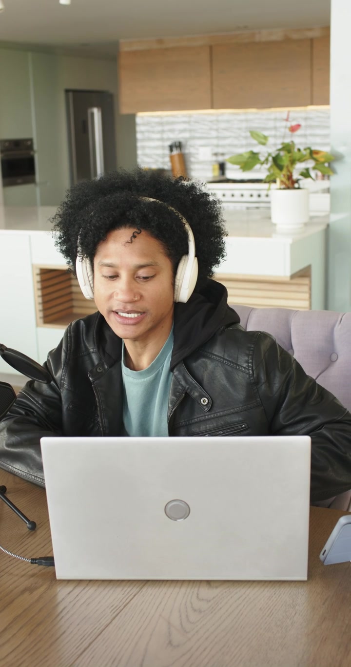 Vertical video: Man sitting in kitchen wearing headphones, speaking into mic, recording on laptop