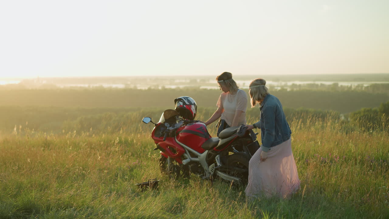 두 여동생은 해가 지는 동안 열린 잔디에서 은 모터사이클을 즐겁게 청소합니다. 한 자매는 옷을 들고 춤을 추고 있습니다.