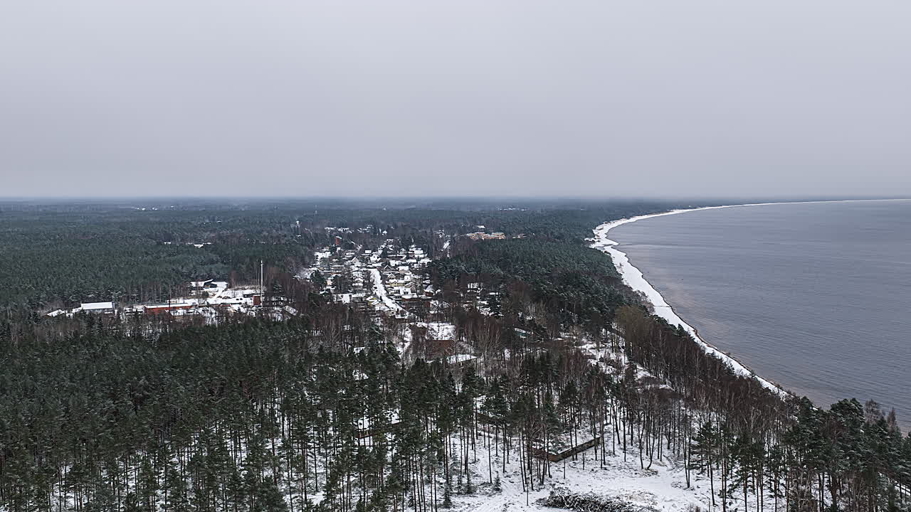 Soft timelapse av skyer fra en drone over snødekt furuskog og en liten kystlandsby, med en lang, hvit strandlinje og et rolig hav som forsvinner inn i vinterdis