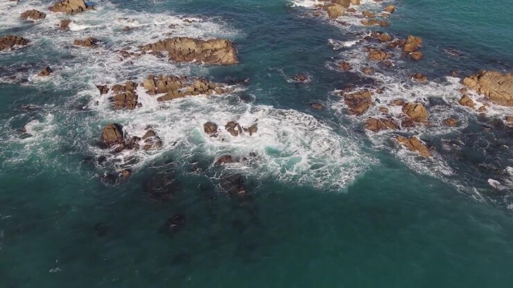 Riprese aeree mozzafiato con drone su scogliere rocciose al sole del tardo pomeriggio, con onde che si infrangono sulle rocce in un mare blu cristallino, cielo azzurro e viste lontane fino all'orizzonte.