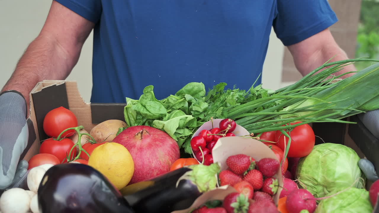 Close up of a man wearing gloves holding a box full of fresh fruits and vegetables