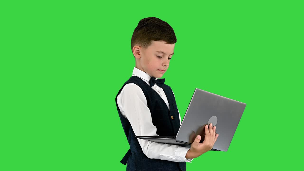 녹색 스크린, 크로마 키에 서 있는 동안 노트북 컴퓨터를 사용하여 타이와 웨스트코트를 입은 웃는 소년