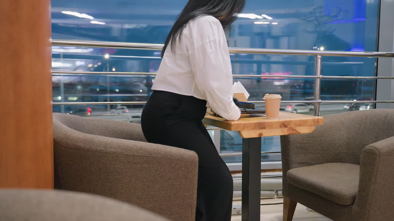 Elegant lady holding drink walks toward cafe table, gently places cup down, sits on chair, and looks through large glass panel reflecting city lights and interior ambiance of modern space