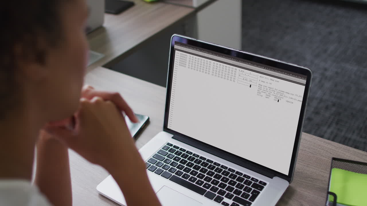 mujer biracial sentada en el escritorio viendo el procesamiento de datos de codificación en la pantalla de la computadora portátil