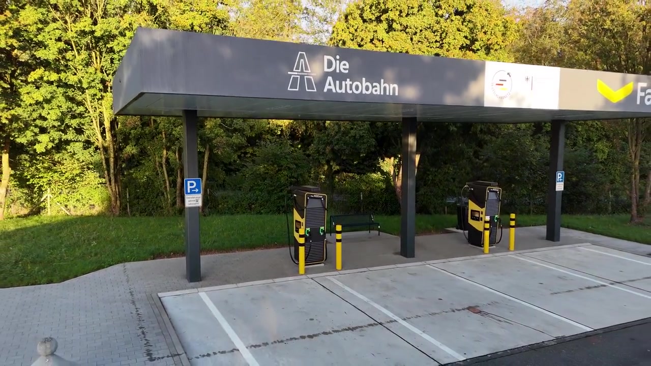 Un lento movimento aereo all'indietro parte vicino a una moderna colonnina di ricarica per veicoli elettrici e rivela l'intera tettoia, la segnaletica visibile, due stazioni di ricarica e l'autostrada vicina