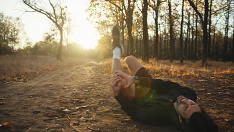 a male athlete in a red cap and black sports uniform holds his knee and falls to the ground during his run because he injured his leg in the morning in the autumn forest at sunrise