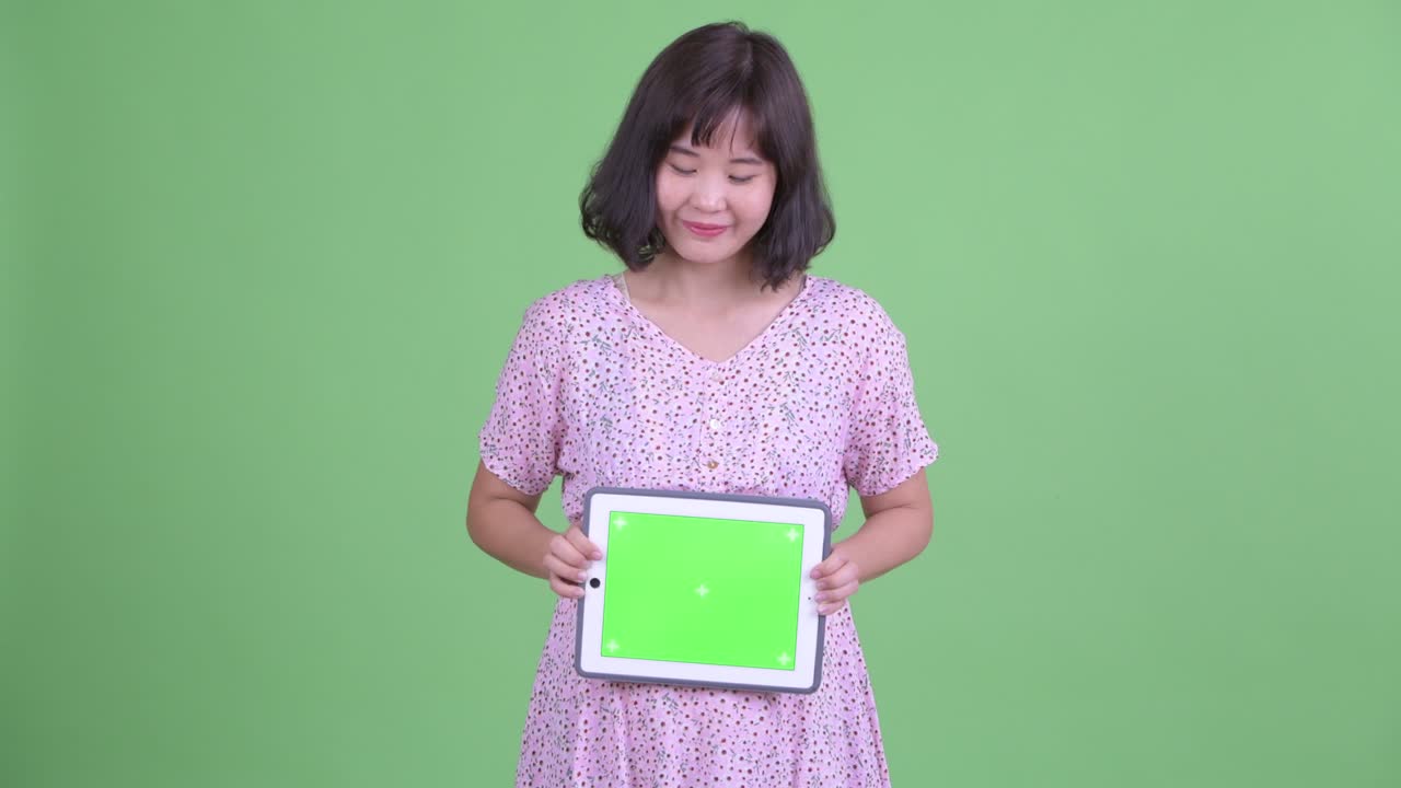 초록색 스튜디오 배경에 미소 짓고 생각하면서 복사 공간이 있는 디지털 태블릿을 들고 있는 아름다운 임신한 아시아 여성의 스튜디오