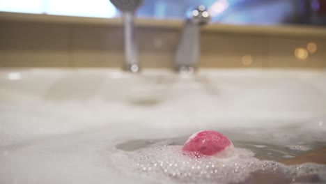 pink bath bomb dissolves in foamy bubbly water in tub, close view