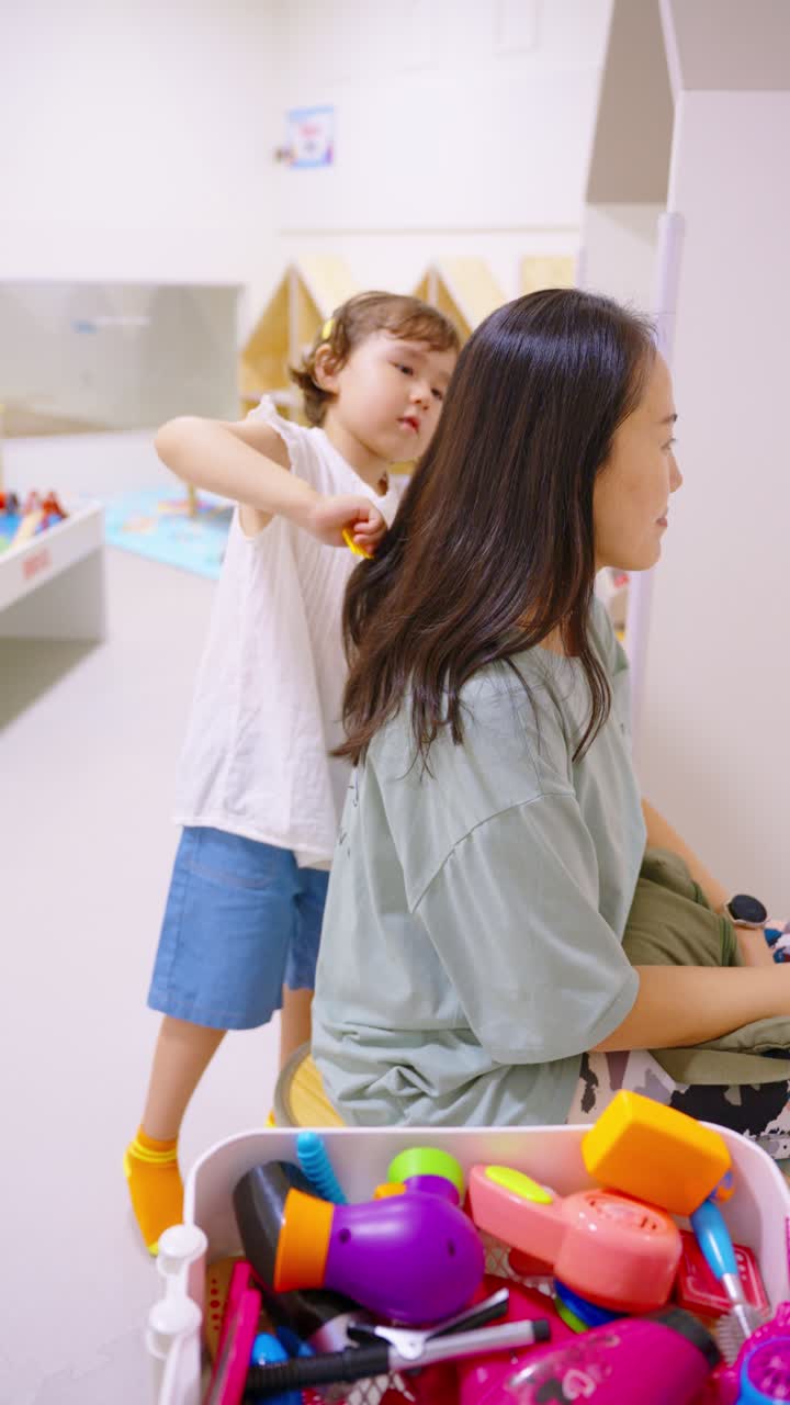 A young girl brushes her mother's hair with a toy comb at an indoor play area, surrounded by colorful toy salon tools