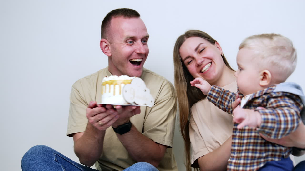 Mother is holding a little baby wearing checkered shirt. Kid tries to reach a cake held by his dad. Family celebration.