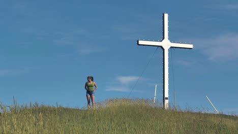 a pretty young woman in shorts and tank top with ponytail stands on top of a hill near a large white cross