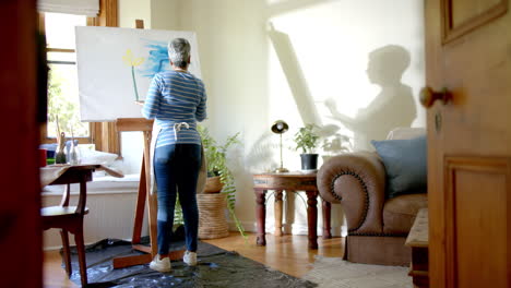 Senior-biracial-woman-wearing-apron-and-painting-on-big-canvas-at-home,-slow-motion