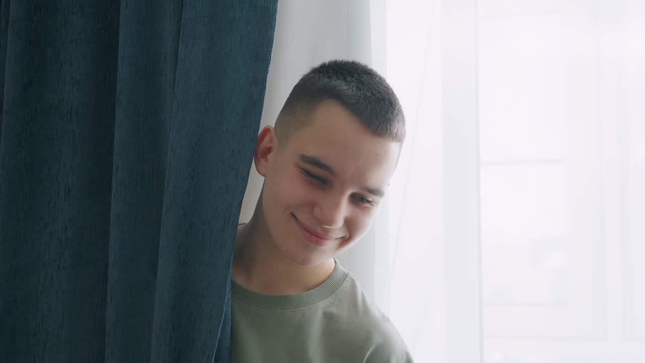 Smiling boy peeks from behind thick dark blue curtain near bright window with sheer white drapes, enjoying quiet playful moment while sunlight softly illuminates calm indoor setting