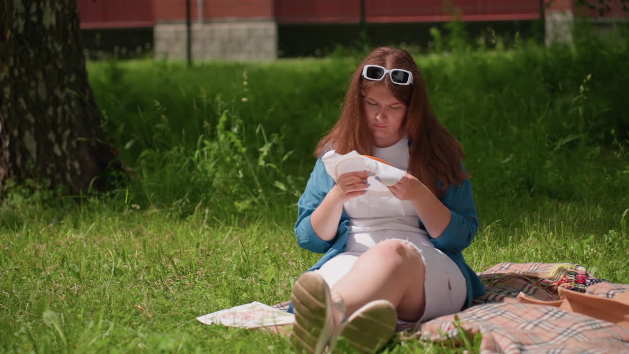 Young girl sitting on blanket under shade of tree in sunny green park working on embroidery design, holding fabric with focus and creativity, surrounded by calm nature