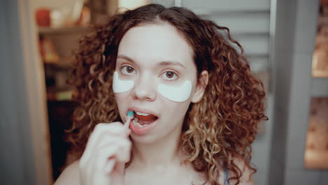 chica de cabello rizado con parches debajo de los ojos cepillándose los dientes en el baño