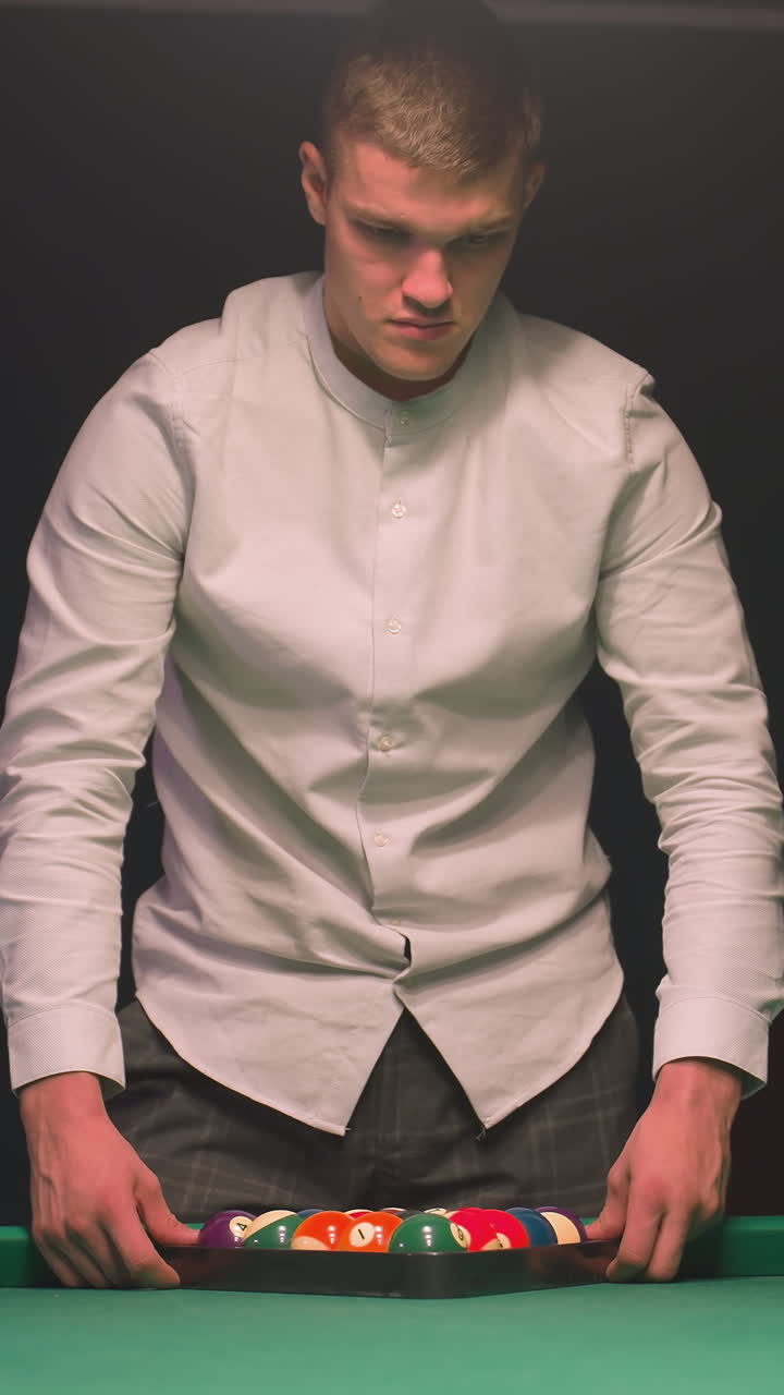 camisa de aprendiz de billar y pantalones grises arregla cuidadosamente bolas de billar de colores en triángulo en la mesa de billar verde. mientras se centra con concentración tranquila en el salón elegante