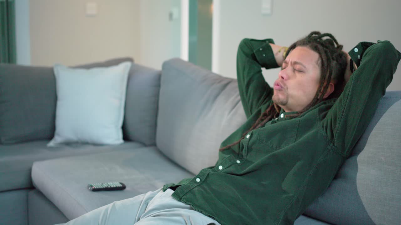Uomo afroamericano sui trentacinque anni mangia popcorn guardando un programma sul divano mentre allunga la mano per prendere il telecomando, spazio per il testo