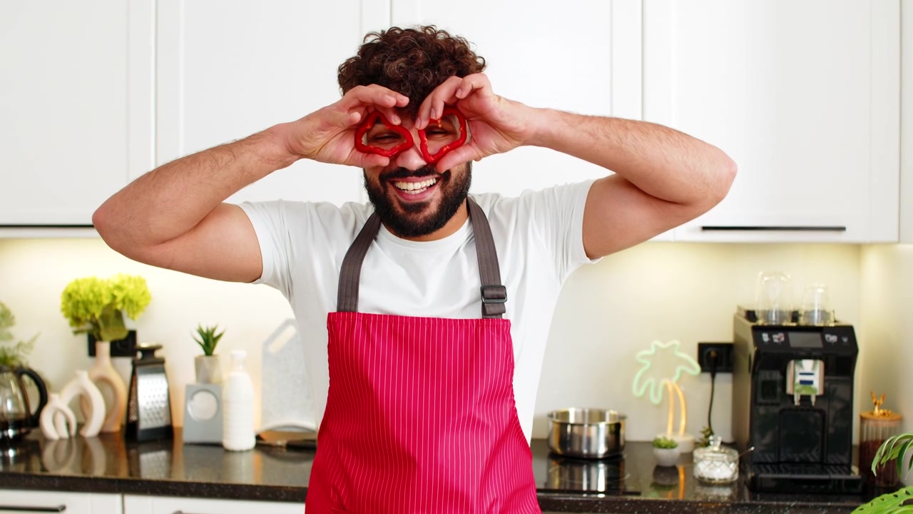 Giovane uomo arabo adulto che scherza in cucina, con anelli di peperone sugli occhi, promuovendo una dieta chetogenica sana