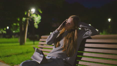 young woman seated on park bench at night, engaged in phone call with laptop on lap, resting head on hand, soft park lighting highlights her focused expression, surrounded by greenery