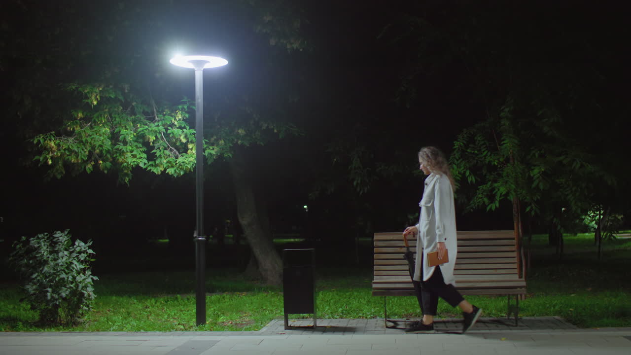 High school student walks to wooden bench under bright streetlight drops umbrella adjusts hair crosses leg opens book to read peacefully while surrounded by trees and calm night atmosphere