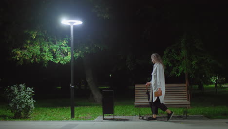 high school student walks to wooden bench under bright streetlight drops umbrella adjusts hair crosses leg opens book to read peacefully while surrounded by trees and calm night atmosphere