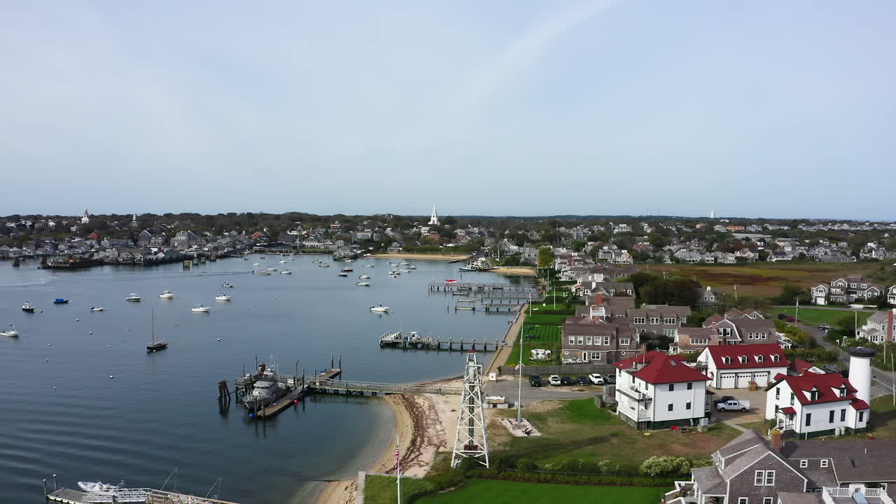 Drone voa para a esquerda sobre Nantucket, Massachusetts, revelando barcos ancorados, píeres em atividade, casas costeiras e distantes torres de igreja que margeiam o porto em uma vista aérea panorâmica da Nova Inglaterra.
