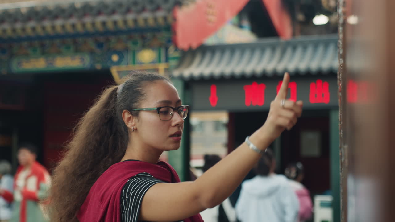 Mujer explorando el patio de un templo chino, llevando bufanda roja y gafas, tocando un pilar de bronce ornamentado con una mirada curiosa, gesto de señalar, retrato espontáneo entre la multitud bulliciosa, techo tradicional.