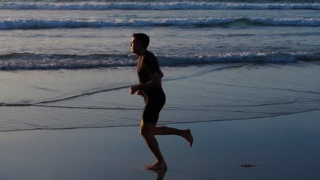 man jogging barefoot on wet sandy beach at sunrise with waves rolling in background. side view, tracking drone shot
