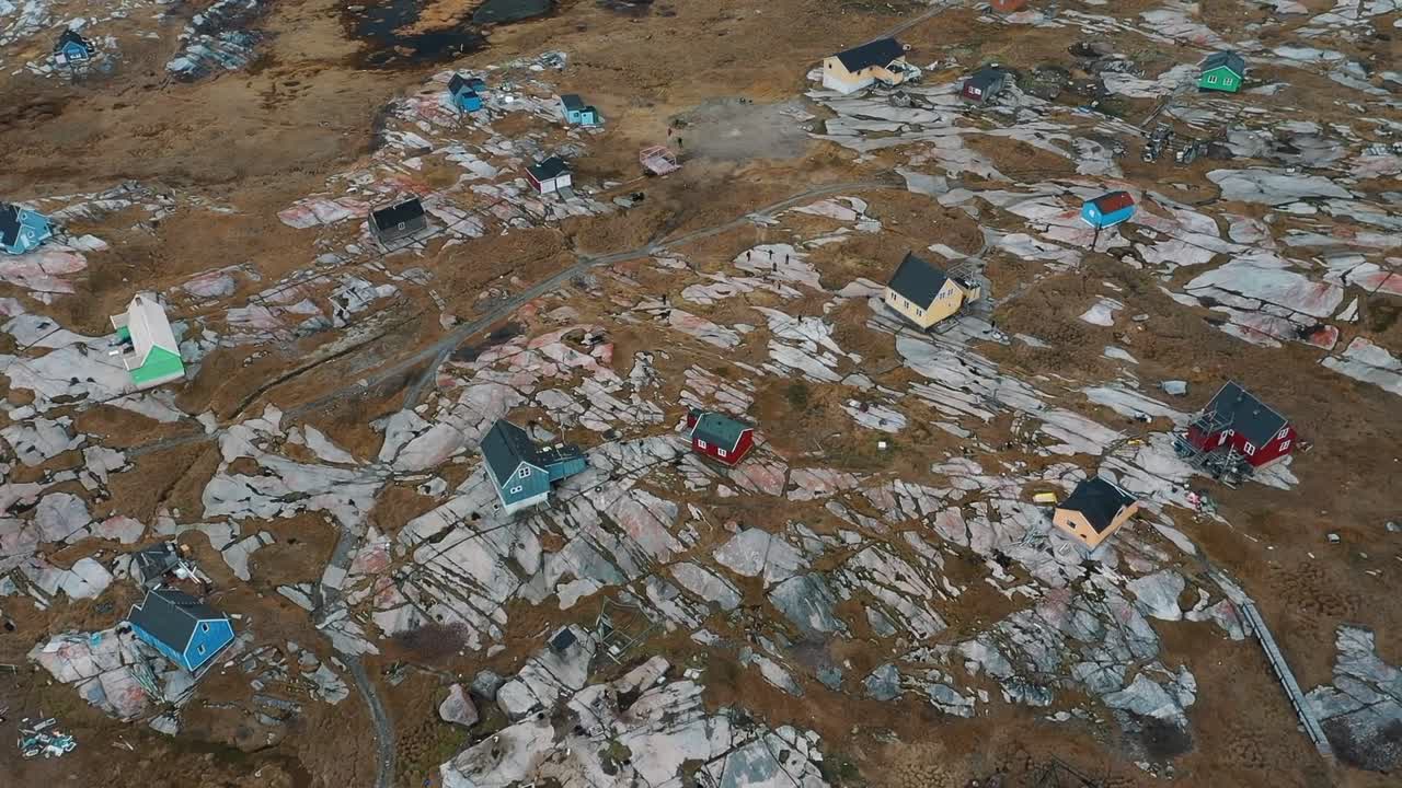 Aerial drone view of colorful wooden houses built along rocky coastline in the remote Arctic village of Ittoqqortoormiit, Greenland, surrounded by clear green water and rugged terrain