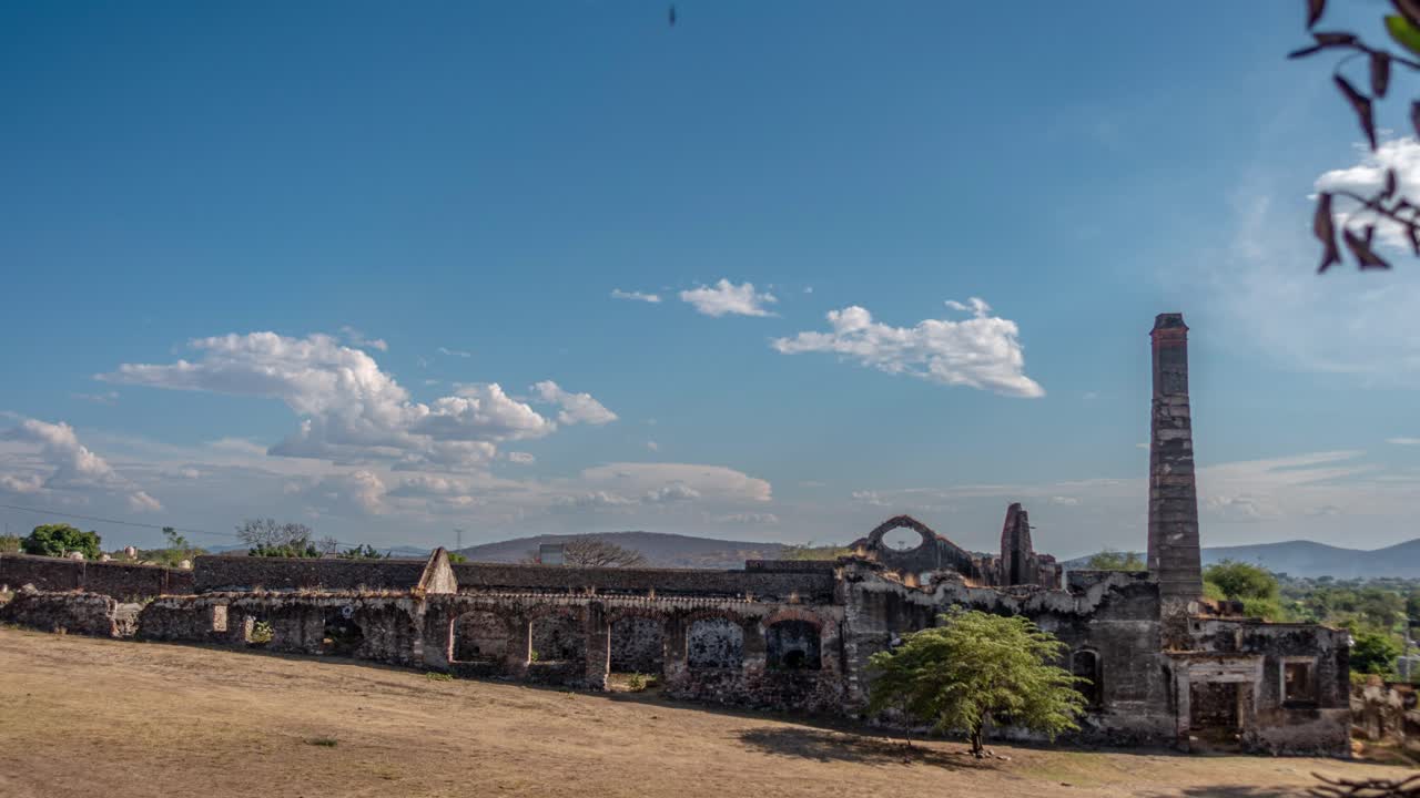 Rovine di un'hacienda storica riprese in time lapse con un alto camino e muri in pietra, paesaggio arido e alberi sparsi sotto un cielo blu che creano un'atmosfera rurale vintage e una scena di patrimonio culturale