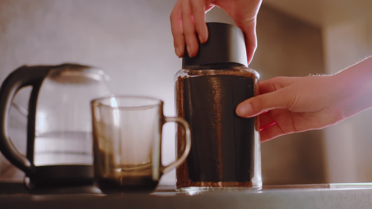 Close view of coffee container placed on countertop beside kettle, hand visible above counter preparing to open lid and scoop granules into cup, natural light highlighting morning routine atmosphere
