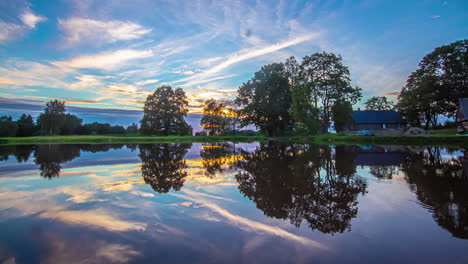 Eine-Farbenfrohe-Wolkenlandschaft-Sonnenuntergang-über-Einem-See-Mit-Einem-Häuschen-Und-Bäumen---Zeitraffer-Mit-Dem-Himmel,-Der-Sich-Auf-Der-Wasseroberfläche-Spiegelt