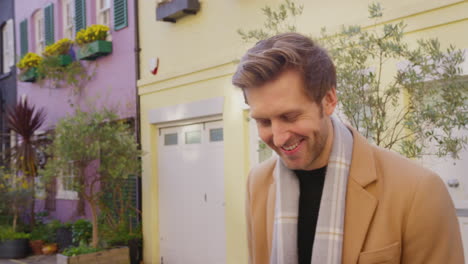 Portrait-of-smiling-man-wearing-coat-and-scarf-standing-on-cobbled-mews-street-on-visit-to-city-in-autumn-or-winter---shot-in-slow-motion