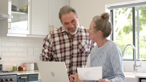 smiling mature couple reviewing documents at home with laptop in kitchen