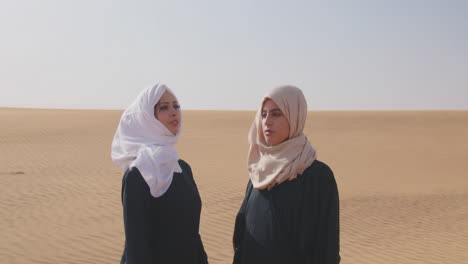 two muslim women wearing traditional dress and hijab standing in a windy desert 1