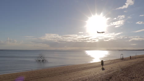 plane approaching airport as it flies over beach and calm sea waters with bright sun shining