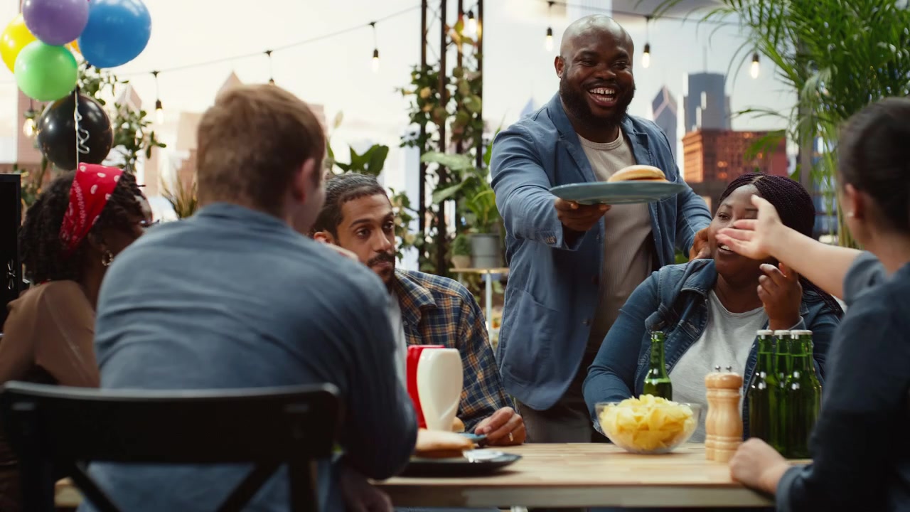 Man serving burger on table from barbecue grill as friends talk and have fun