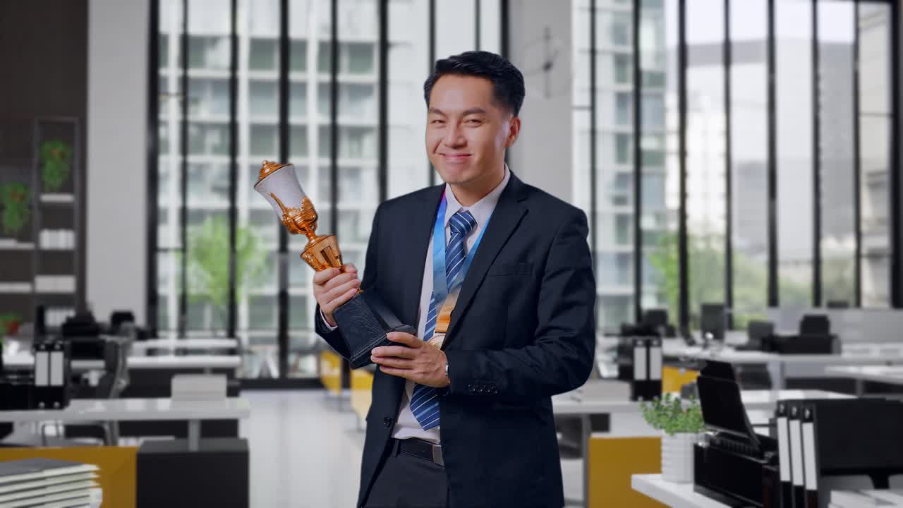 Doanh nhân châu Á mặc vest và cà vạt, với huy chương vàng và cúp, đang hét lên ăn mừng chiến thắng với tư cách là người chiến thắng đầu tiên tại văn phòng.
