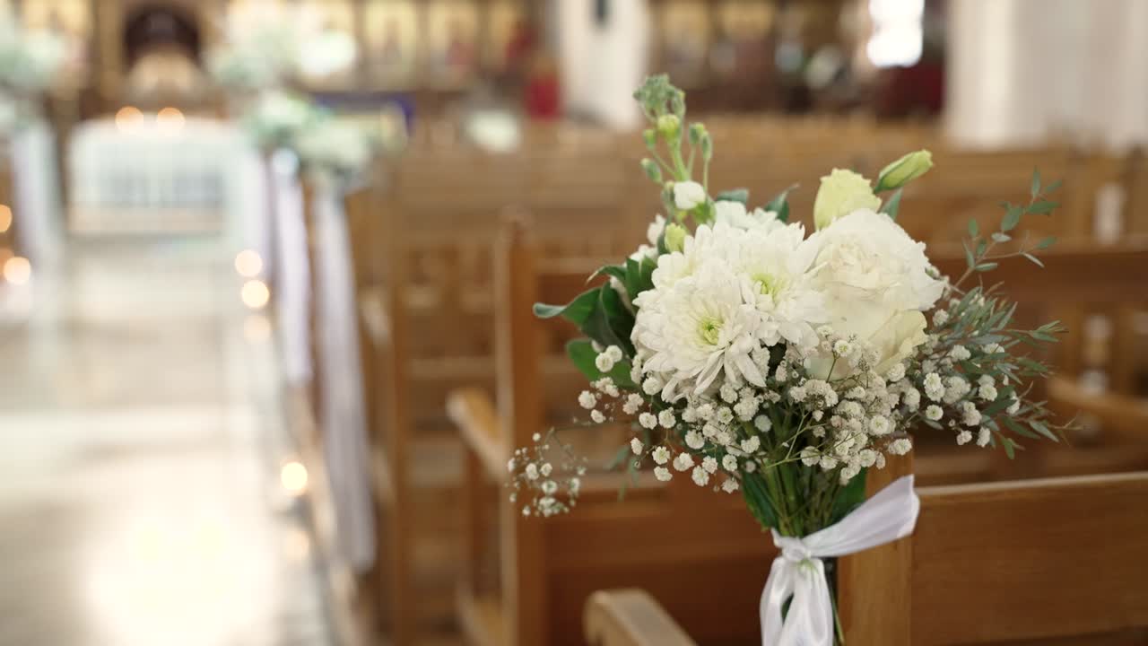 Fiori bianchi sono disposti sulle sedie in una chiesa. L'ambiente è preparato per una cerimonia nuziale. Gli eventi si svolgono nel pomeriggio con una luce soffusa.
