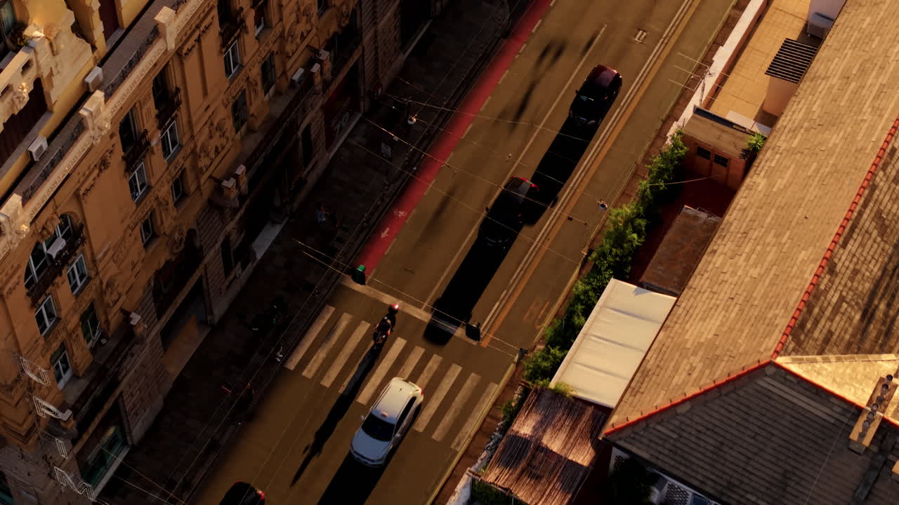 Drone starts above rooftops in Genoa, tilts down into street view with cars and scooter, ending in a full top-down view at sunset
