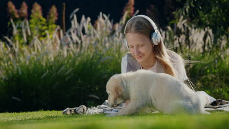 Teenage-girl-plays-with-a-puppy,-lies-on-the-lawn,-listens-to-music-on-headphones