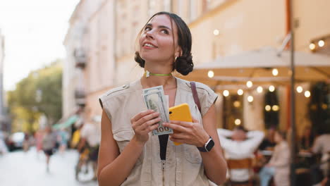 Happy-young-woman-counting-money-dollar-cash,-use-smartphone-calculator-app-in-urban-city-street