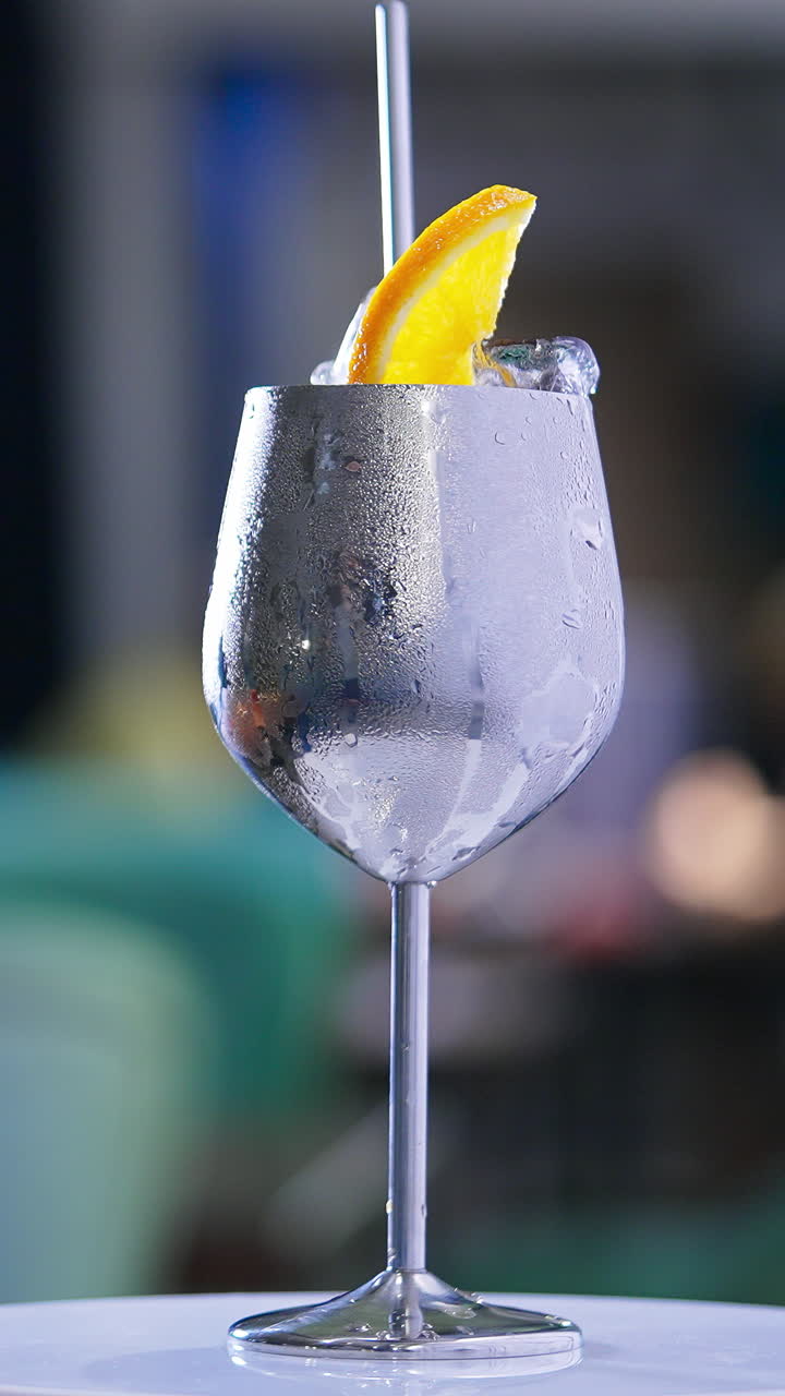Misted metal wineglass with metal straw inside on the rotating table. Cocktail with ice cubes and orange slice on top. Close up. Blurred backdrop. Vertical video