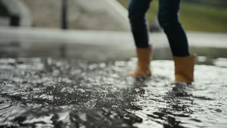 a teenager in orange boots actively walks through a large puddle and pushes the water from side to side on the street during the rain