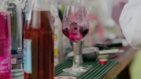 bartender pouring berry cocktail with pink liquid and fruits into glass at bar with colorful bottles