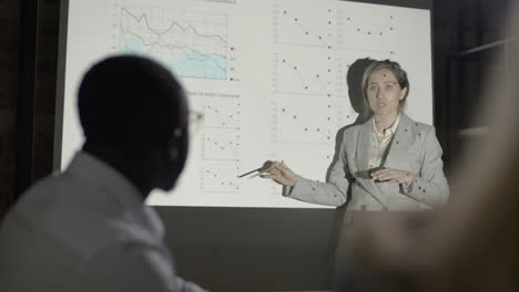 female employee explaning a slide with graphics to two coworkers who are sitting at table in a dark room 1