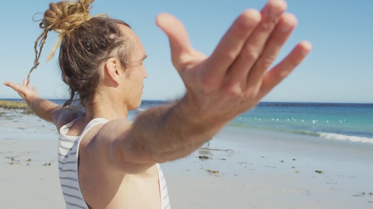 Video di un uomo caucasico con dreadlock che pratica yoga su una spiaggia assolata con le braccia distese.