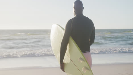 Feliz-Hombre-Afroamericano-Mayor-Caminando-Y-Sosteniendo-Tabla-De-Surf-En-La-Playa,-En-Cámara-Lenta